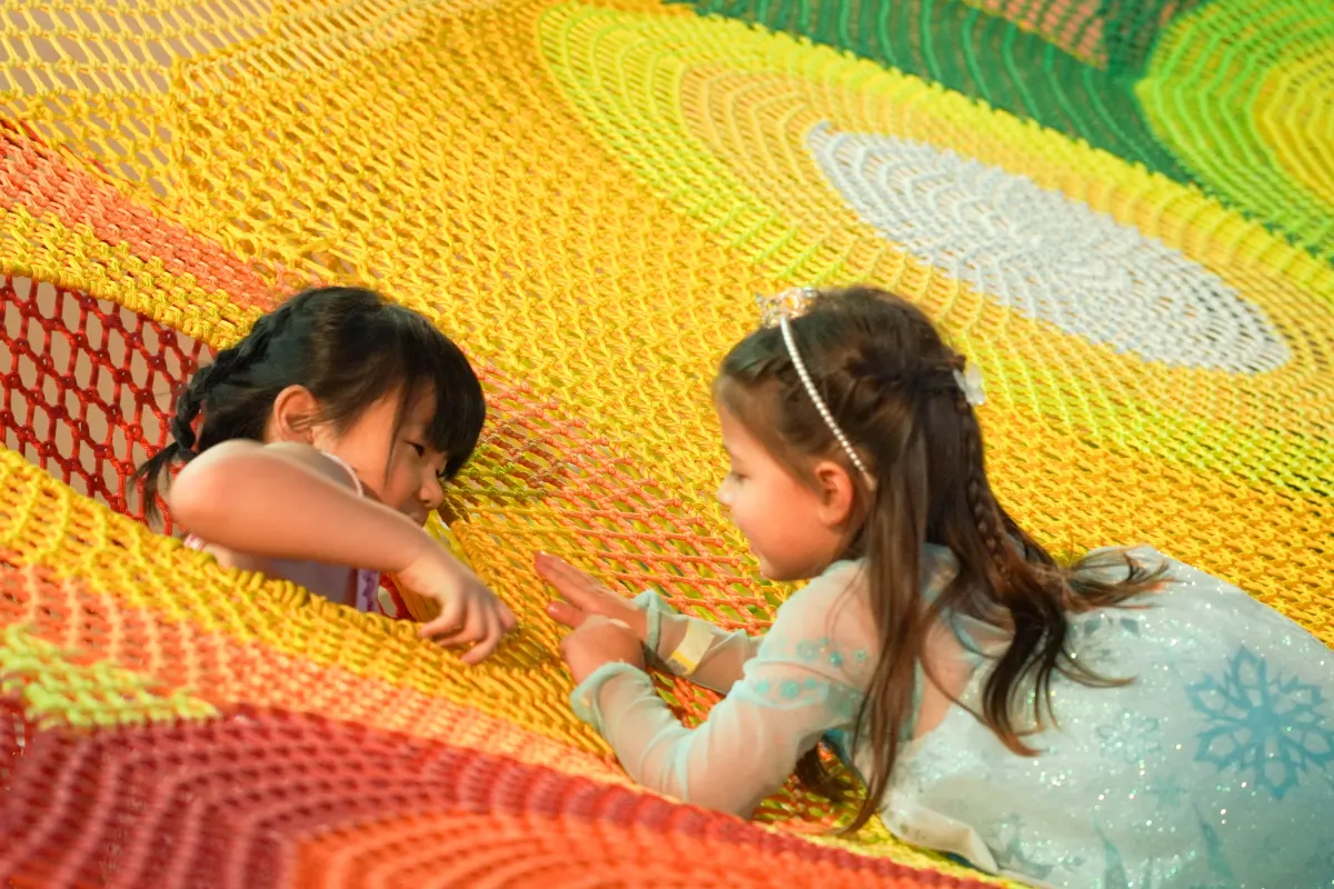 Two girls lie face to face on the giant rainbow crocheted climbing net in the Adventure Trails zone at UFOREA Penang