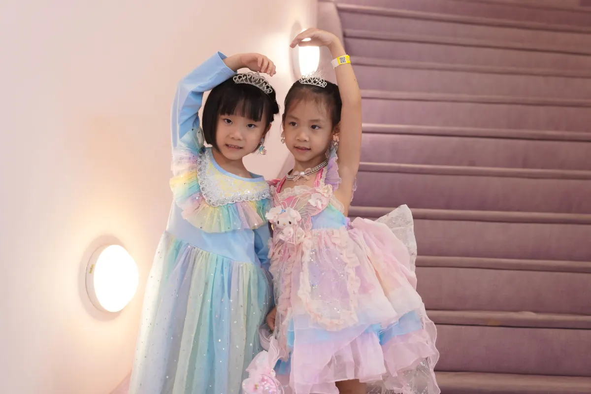 Two girls in rainbow tulle gowns and tiaras arch their arms overhead on the lilac Makeover Castle staircase at UFOREA Penang