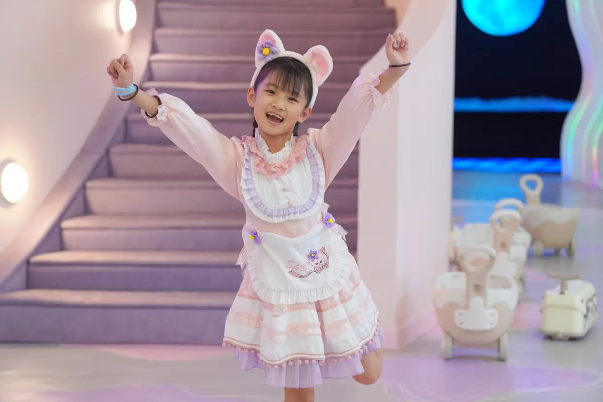 A girl in a pastel cat-eared pinafore dress throws both arms up in delight at the foot of the lilac Makeover Castle staircase at UFOREA Penang