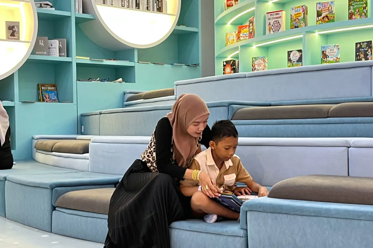 A mother in a hijab reads a picture book with her son on the tiered blue seating of the Cozy Nook, framed by round backlit bookshelves, at UFOREA Penang