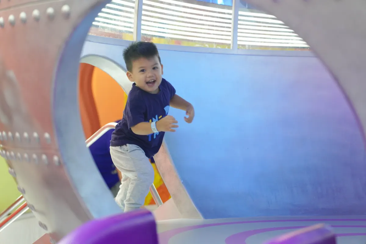 A boy laughs as he climbs through the riveted porthole of a blue and purple soft-play tunnel at UFOREA Penang