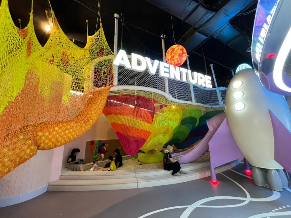 The Adventure Trails zone at UFOREA Penang, a dramatic multi-storey rainbow climbing-net structure with a glowing planet sign, a curving slide and a rocket-shaped tower over a sandy play pit where children climb and explore.