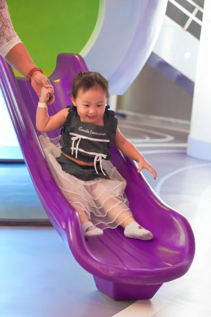 A delighted toddler in a black top and white tutu slides down the purple planet slide, holding a parent's hand, on the lavender space-track floor of the Galaxy Exploration zone at UFOREA Penang.
