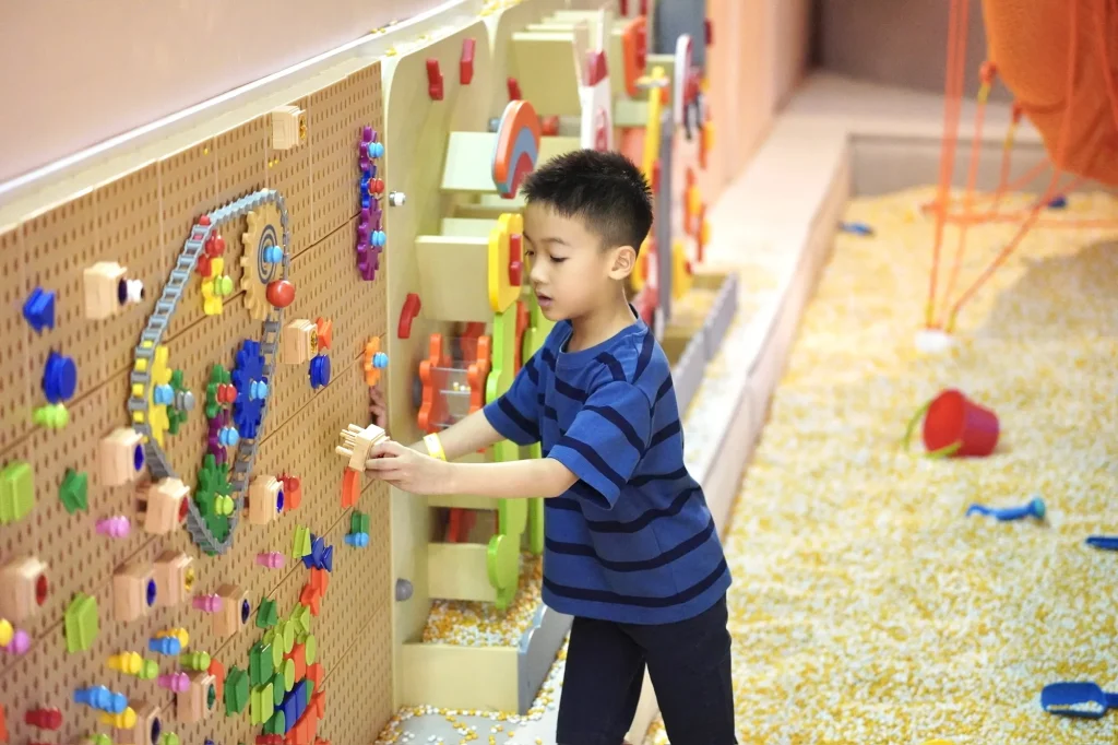 A young boy in a navy striped tee builds with colourful wooden pegs on the busy-board activity wall beside the kernel sand pit in the Adventure Trails zone at UFOREA Penang.
