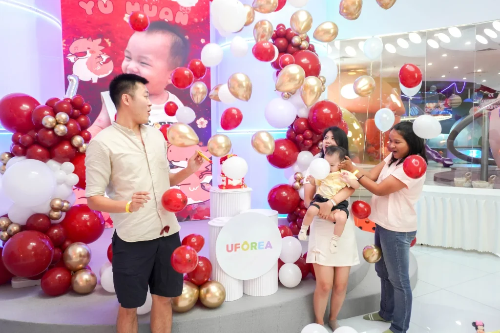 A family celebrates a child's birthday at UFOREA Penang in front of a red, gold and white balloon arch with a personalised baby photo backdrop, while a parent holds a smiling toddler beside the UFOREA-branded party props.