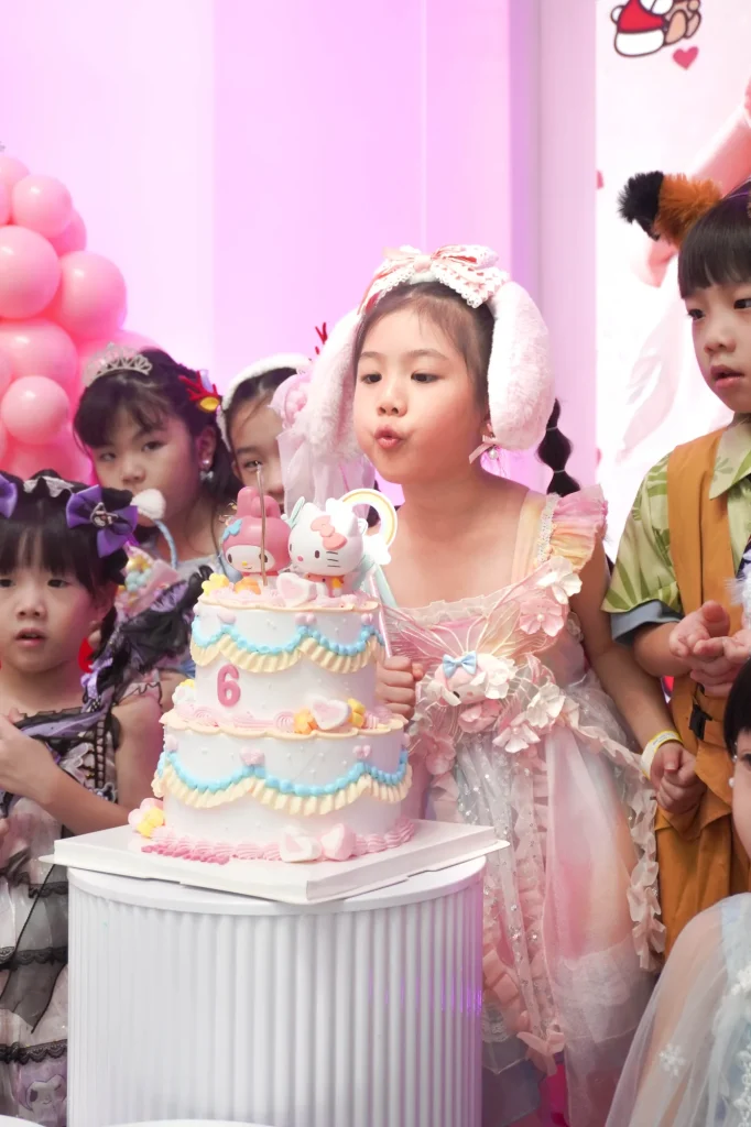 A young girl in a pink princess dress blows out the candle on her two-tier Hello Kitty themed birthday cake as friends look on at a UFOREA Penang birthday celebration framed by soft pink balloons.