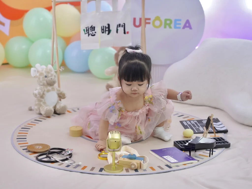 A toddler in a pink tulle dress crawls across a pastel play mat reaching for golden role-play props, a stethoscope, microphone and toy piano, at a first-birthday celebration at UFOREA Penang with the rainbow UFOREA logo behind her.