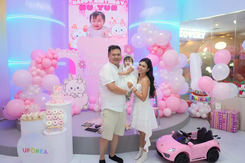 Parents hold their one-year-old in front of a soft-pink balloon arch and 'Happy Birthday Hu Yun' backdrop at UFOREA Penang, beside a dessert table and a pink toy ride-on car, with the Discovery Cove zone sign visible at right.