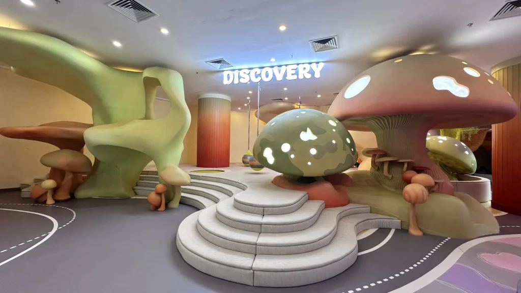 A whimsical sculpted mushroom-forest play landscape at UFOREA Penang with oversized soft green and red toadstools, flowing organic climbing forms and tiered cushioned steps under a glowing 'DISCOVERY' sign.