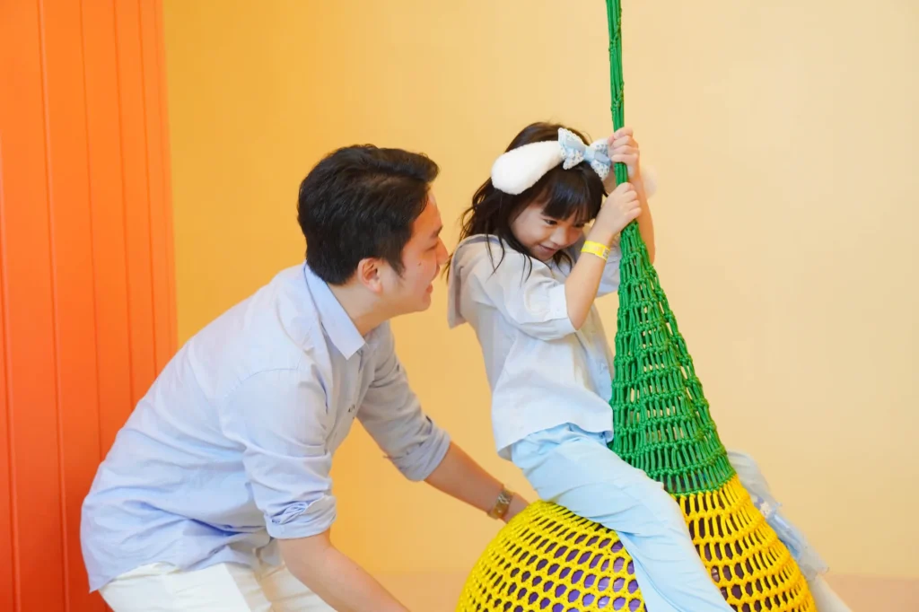 A smiling father gently steadies his little daughter, who wears a white hair bow, as she sits on a woven green-and-yellow rope swing ball against warm orange walls at UFOREA Penang.