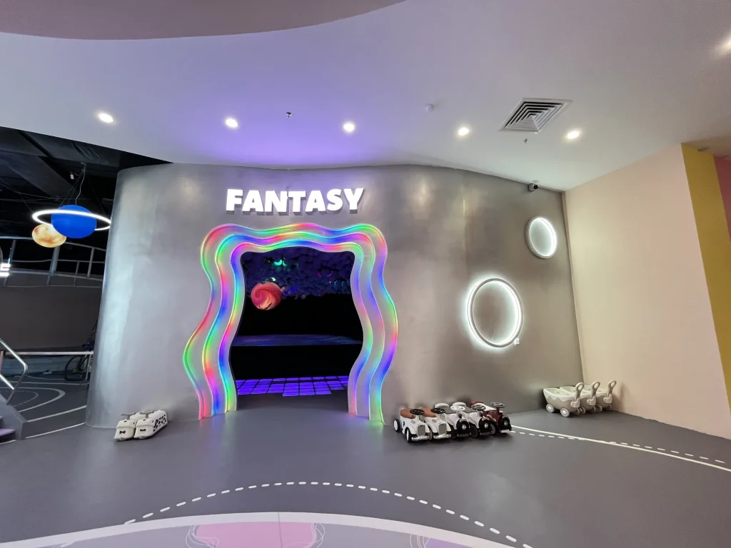 The Dazzling Light Fantasy zone entrance at UFOREA Penang, a glowing rainbow LED archway marked 'FANTASY' leading into a dark LED-lit ball pit, with floating planet decorations and ride-on toy cars parked nearby.