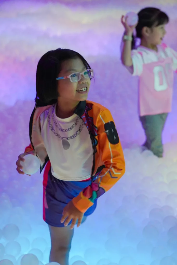 A smiling girl in glasses holds white play balls amid the glowing lavender and pink LED-lit ball pit at UFOREA Penang's Dazzling Light Fantasy zone
