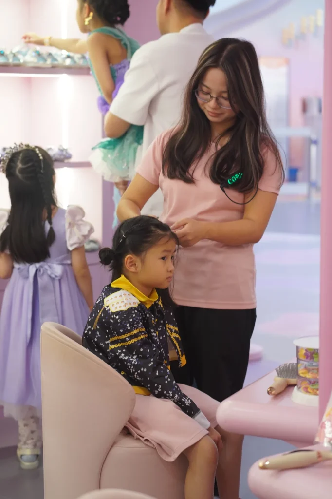 A stylist gently styles a young guest's hair in the coral-pink princess salon at UFOREA Penang's Makeover Castle zone, with other children in dress-up gowns nearby