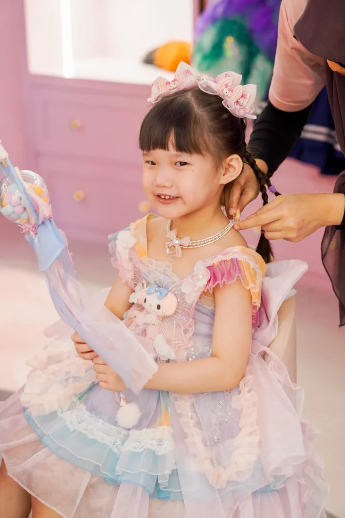 A smiling young girl in a pastel layered princess dress and a sparkly pink bow has her hair braided by a stylist at the dress-up and salon area of the Makeover Castle zone at UFOREA Penang.