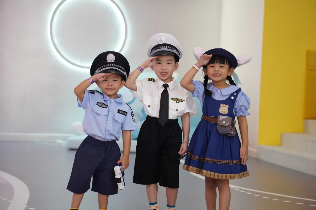 Three young children dressed in role-play police, pilot and uniform costumes stand saluting together in the role-play area at UFOREA Penang, smiling in front of a clean white set.