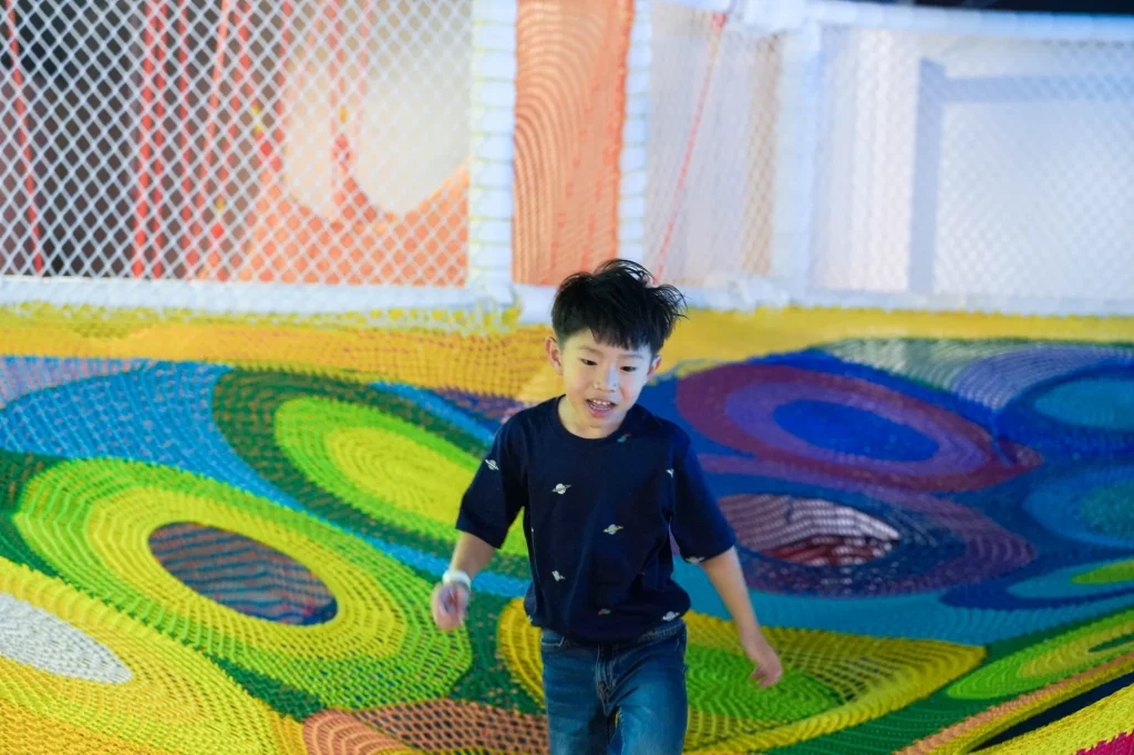 A boy runs and plays across the colorful peacock-patterned trampoline climbing net inside UFOREA Penang's Adventure Trails zone
