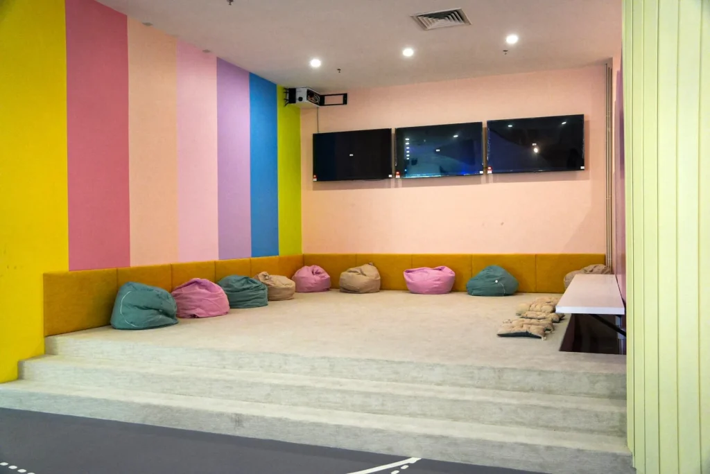 The parent rest and viewing area at UFOREA Penang, a carpeted tiered platform scattered with pastel pink and teal beanbags against a rainbow-striped wall, with three wall-mounted screens above for relaxing while children play.
