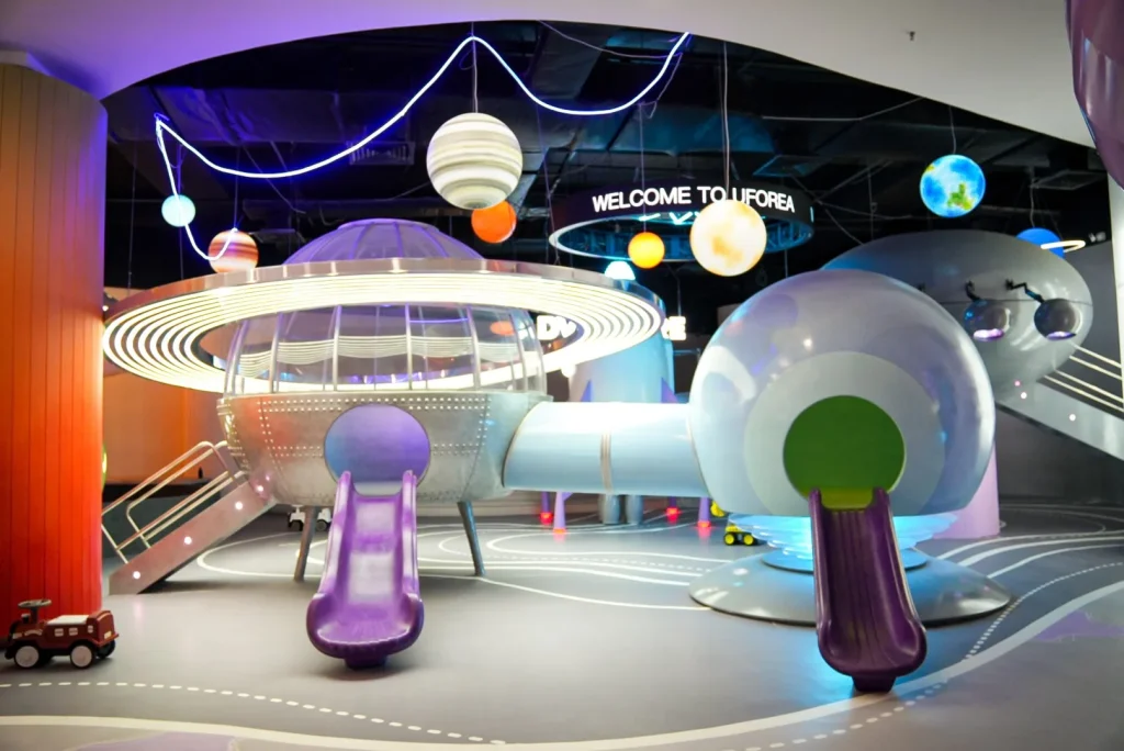 The Galaxy Exploration zone at UFOREA Penang, featuring a giant silver flying-saucer play structure with twin purple spiral slides, hanging illuminated planets and a 'WELCOME TO UFOREA' sign glowing under deep-blue space lighting.