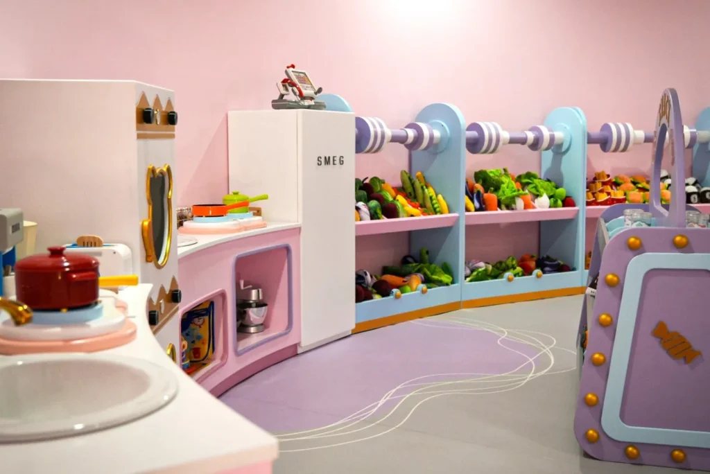 Pastel-pink play kitchen and grocery role-play corner in UFOREA Penang's Mini Town zone, with a toy SMEG fridge, miniature stove and red pot, and bins of plush fruit and vegetables on candy-striped shelves.
