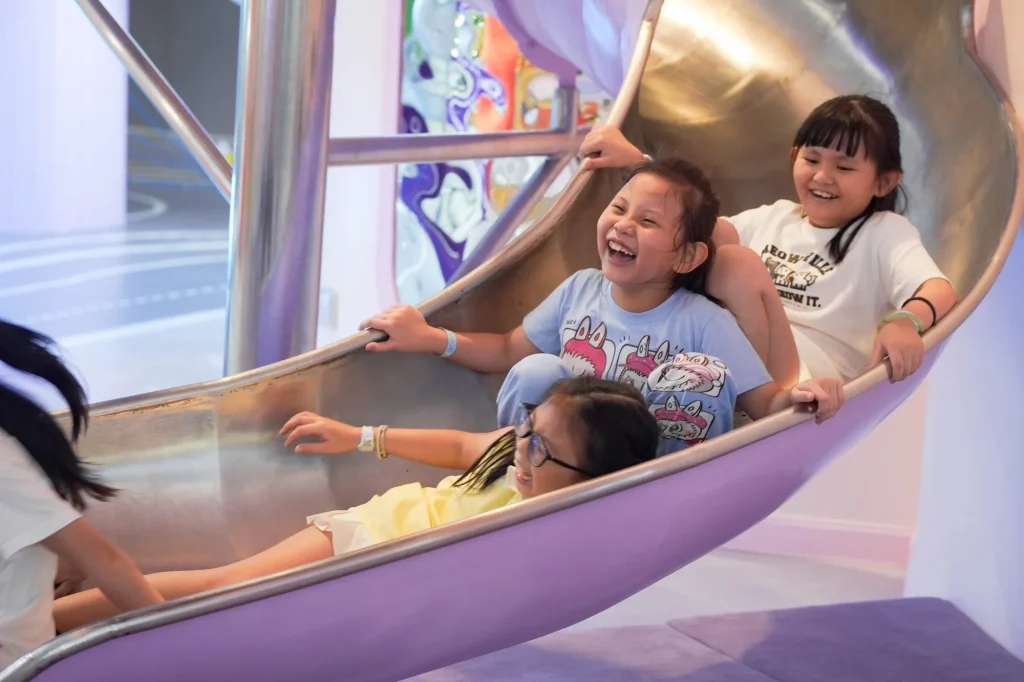 Three girls laugh with delight as they ride down the curved metallic space slide in the purple-themed Galaxy Exploration zone at UFOREA Penang